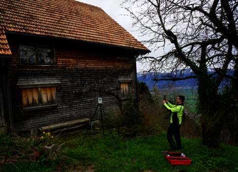 Eine Person in einer leuchtend gelben Jacke positioniert eine Kamera auf einem Stativ neben einem alten, holzverkleideten Haus mit rotem Ziegeldach; ein roter Werkzeugkasten liegt auf dem Gras, im Hintergrund eine ländliche Landschaft mit kahlen Bäumen.