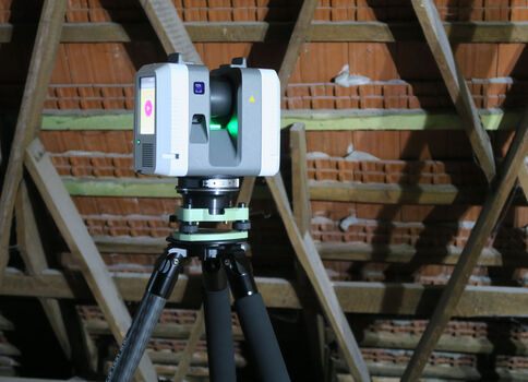 Vermessungsgerät (Laserscanner) auf einem Stativ; steht auf einer Baustelle, im Hintergrund sind Holzbalken des Dachstuhls und eine Backsteinwand sichtbar.