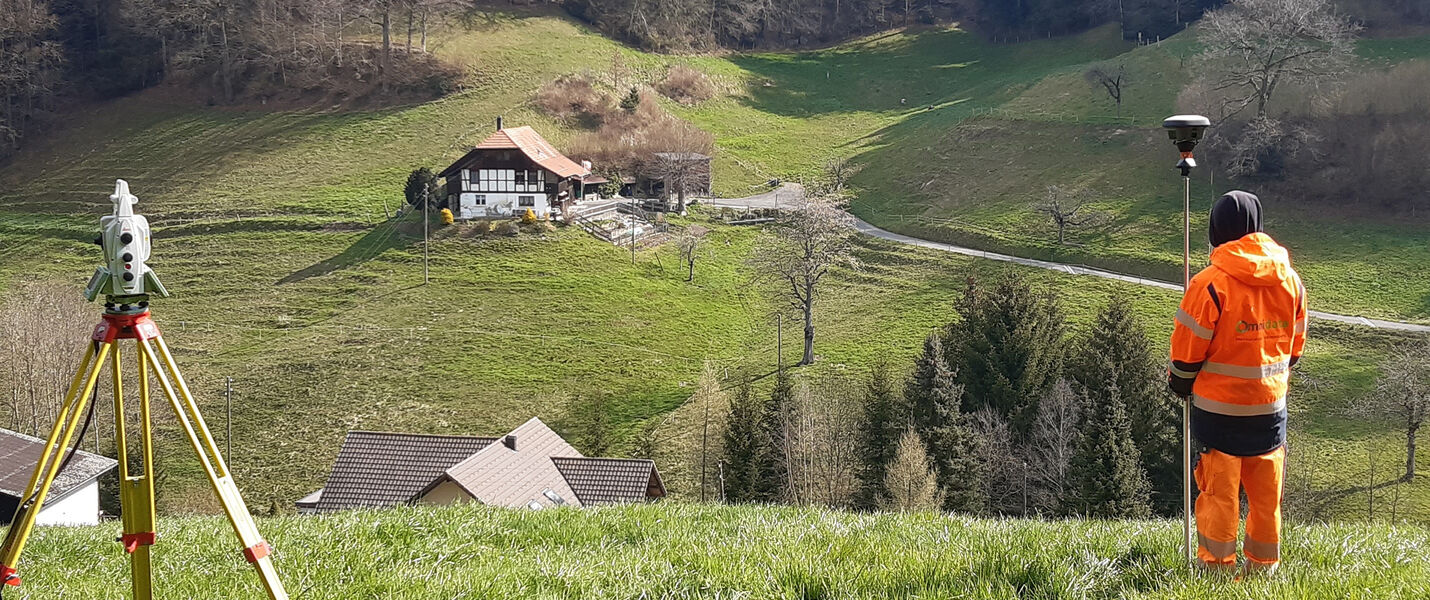 Un agent de terrain en tenue de sécurité orange se tient près d’un trépied doté d’un instrument de topographie, dans un paysage rural vallonné avec quelques maisons au loin.