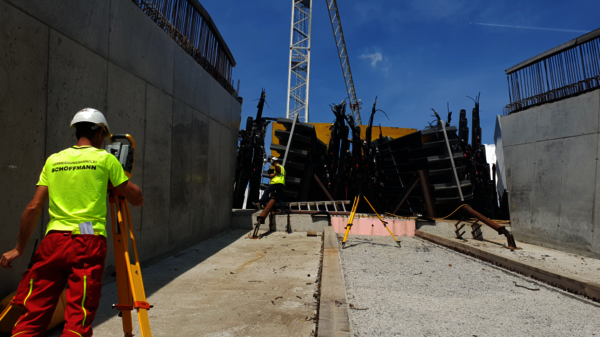 Two construction workers in high-visibility clothing and hard hats operate at a site between concrete walls. One uses a tripod-mounted instrument near a railing while a crane and a pile of steel beams lie behind them on railway tracks.