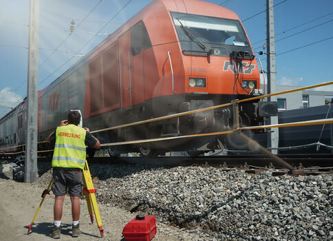 Eine Person in fluoreszierender Warnweste steht an Bahngleisen neben einem roten Lokzug und bedient eine Vermessungsapparatur; ein roter Werkzeugkoffer liegt auf dem Kies, im Hintergrund sind Oberleitungen und ein zweiter Zug zu sehen.