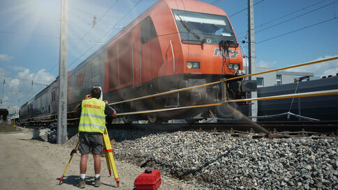 Eine Person in fluoreszierender Warnweste steht an Bahngleisen neben einem roten Lokzug und bedient eine Vermessungsapparatur; ein roter Werkzeugkoffer liegt auf dem Kies, im Hintergrund sind Oberleitungen und ein zweiter Zug zu sehen.