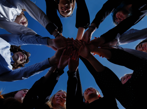 Eine Gruppe von Menschen steht im Kreis und legt ihre Hände gemeinsam in die Mitte übereinander – Symbol für Zusammenarbeit und Gemeinschaft vor blauem Himmel.