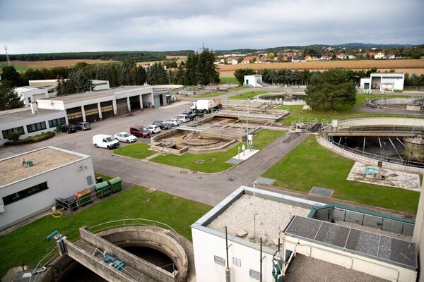 Luftaufnahme eines Abwasserreinigungswerks: Mehrere runde Becken, weiße Betongebäude, Parkplätze mit Autos, Wege und Grünflächen; im Hintergrund weitere Anlagen und eine ländliche Umgebung.