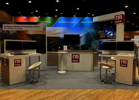 Ein Messestand von RM DATA mit drei Arbeitsstationen (Monitore) und Barhockern. Zentraler Empfangstresen mit RM DATA-Logo; farbige Banner im Hintergrund thematisieren IT-Begriffe wie Datenmanagement und Reality Capture.