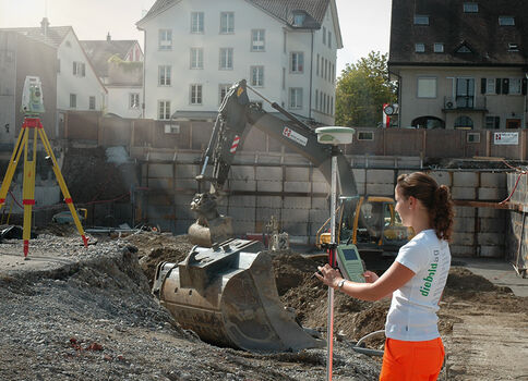 Auf einer Baustelle führt eine Person Vermessungsarbeiten mit einem Messgerät durch; im Hintergrund arbeitet ein Bagger, während Absperrungen und Wohnhäuser das Gelände säumen.