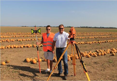 Zwei Männer stehen in einem Kürbisfeld; einer trägt eine orange Warnweste, der andere ein weißes Hemd. Zwischen ihnen stehen Vermessungsgeräte auf Stativ, während orangefarbene Kürbisse die Felder im Hintergrund säumen.