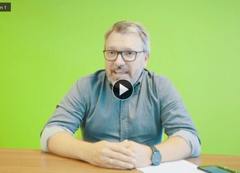 Ein Mann mit Brille und kurzer grauer Bart sitzt an einem Holztisch vor einem leuchtend grünen Hintergrund. Er schaut in die Kamera, seine Hände liegen gefaltet vor ihm; auf dem Tisch liegen Papier und ein Smartphone, während im Bild ein Video-Play-Symbol sichtbar ist.