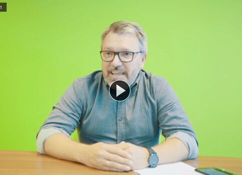 Un homme portant des lunettes et une barbe est assis à un bureau devant un fond vert vif; il parle dans une vidéo, avec des feuilles et un téléphone sur le bureau et une icône de lecture au centre.