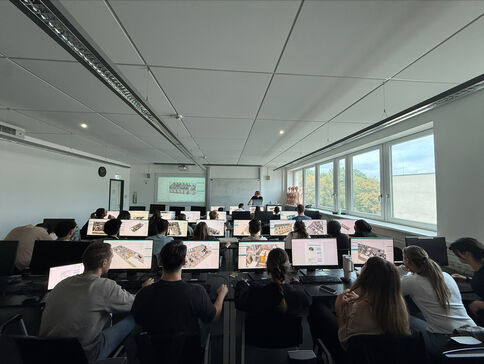 Eine Lehrveranstaltung in einem Computerraum: Dozent steht vorne neben einer präsentierten Folie, Studierende sitzen an Computerarbeitsplätzen in Reihen, rechts großes Fenster mit Blick nach draußen.
