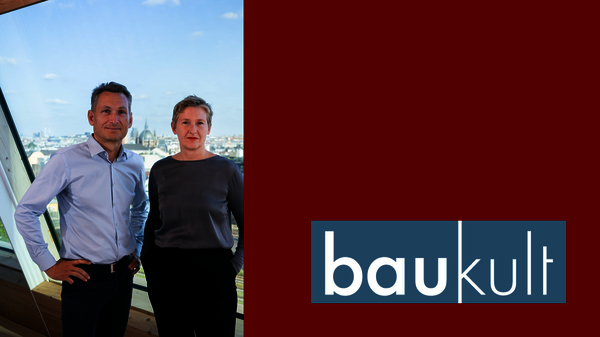 Zwei Personen, ein Mann und eine Frau, posieren in einem Innenraum vor einem Fenster mit Stadtblick; rechts ein Logo "baukult" auf dunkelrotem Hintergrund.