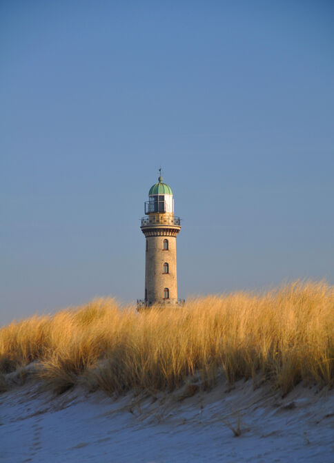 Ein Steinleuchtturm steht auf Dünen, von goldenem Gras umgeben; der klare blaue Himmel bildet den Hintergrund.