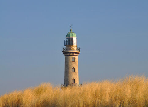 Ein Steinleuchtturm steht auf Dünen, von goldenem Gras umgeben; der klare blaue Himmel bildet den Hintergrund.