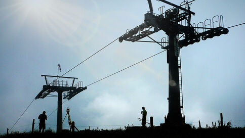 Überwachungsmessung Zwei große Stahl-Türme einer Seilbahn- oder Skiliftanlage tragen ein gespanntes Drahtseil; am Hang stehen einige Personen, während der Himmel blau und sonnig ist.