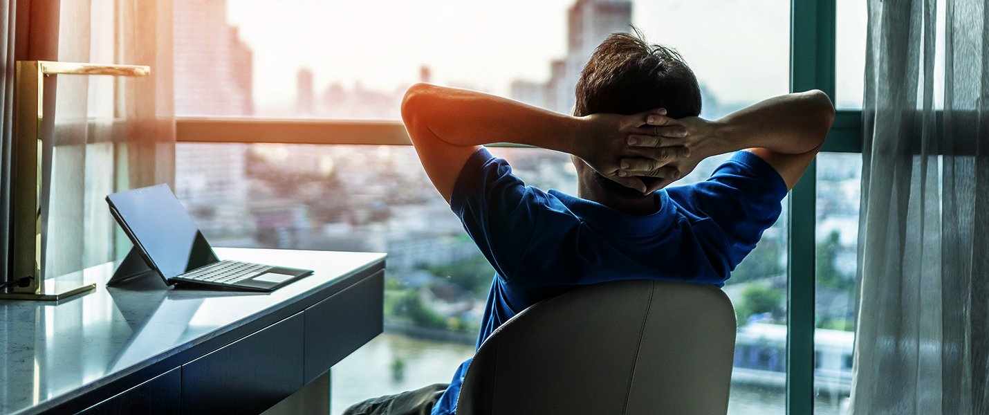 Ein Mann sitzt entspannt an einem Arbeitsplatz, die Hände hinter dem Kopf verschränkt, Blick auf eine Stadt durch ein großes Fenster. Auf dem Schreibtisch liegen Laptop und Tablet; warmes Tageslicht beleuchtet den Raum.
