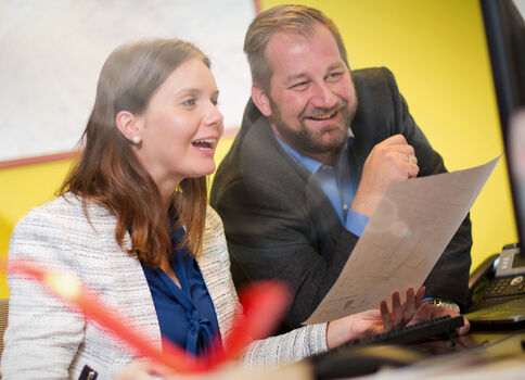 Zwei Kollegen sitzen an einem Schreibtisch im Büro, schauen gemeinsam auf ein Blatt Papier und lachen. Im Vordergrund ist ein Computerbildschirm zu sehen; im Hintergrund eine gelbe Wand.