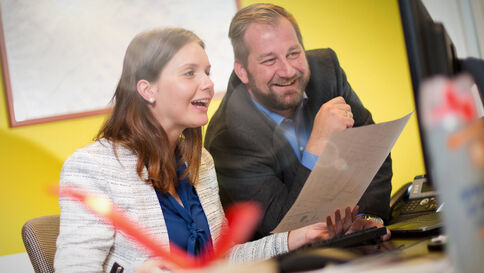Zwei Kollegen sitzen an einem Schreibtisch im Büro, schauen gemeinsam auf ein Blatt Papier und lachen. Im Vordergrund ist ein Computerbildschirm zu sehen; im Hintergrund eine gelbe Wand.