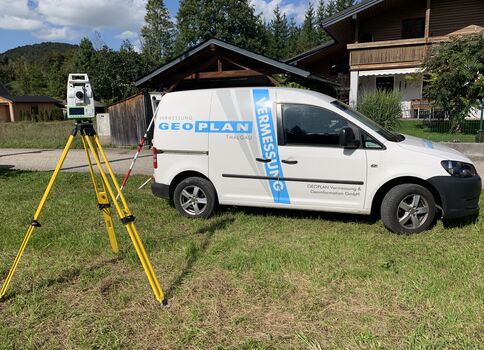 Weißer Vermessungswagen mit blauer Beschriftung vor einem Haus auf grünem Rasen; davor ein gelbes Stativ mit Vermessungsgerät. Hintergrund: Wald und Häuser.