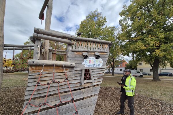 Ein hölzernes Spielschiff auf einem Spielplatz, mit einem Seilnetz an der Seite. Ein Mann in leuchtend gelber Warnweste steht daneben und hält einen Block; im Hintergrund Bäume und Häuser.