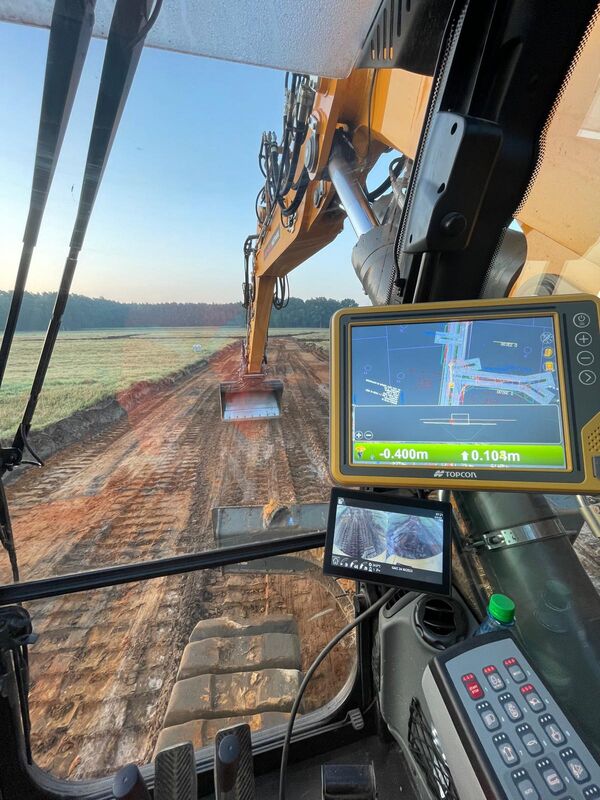 View from inside a heavy excavator cabin as the yellow hydraulic arm extends forward to dig a dirt trench. An onboard monitor shows a diagram and distance readings. Outside, a dirt track leads to a field and distant treeline under a clear blue sky.