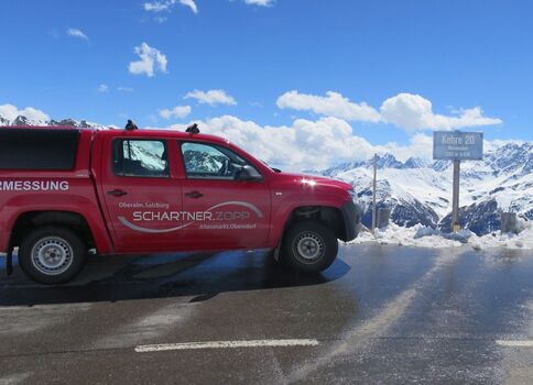 Rotes Vermessungsfahrzeug auf einer schneebedeckten Bergstraße, vor majestätischen schneebedeckten Bergen; ein Straßenschild steht am Straßenrand.