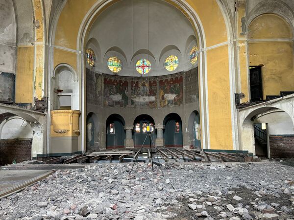 Innenansicht einer zerstörten Kirche: Trümmer bedecken den Boden, zerfallene Wände und Bögen, bunte Glasfenster und Fresken an der Apsis, ein Stativ steht in der Mitte.
