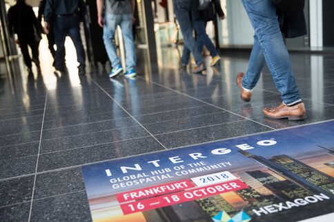 Mehrere Passanten gehen durch eine Halle; auf dem Boden liegt ein großes Poster, das INTERGEO bewirbt – Frankfurt, 16.–18. Oktober 2018 – mit Logos im Hintergrund.