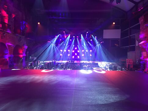 Große Indoor-Bühne in dunklem Saal, von blauen und lila Scheinwerfern beleuchtet. Auf der Bühne stehen Ausrüstung und Aufbau; im Vordergrund eine leere Tanzfläche, über die breite Lichtstrahlen nach unten ziehen.
