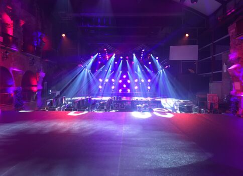 Große Indoor-Bühne in dunklem Saal, von blauen und lila Scheinwerfern beleuchtet. Auf der Bühne stehen Ausrüstung und Aufbau; im Vordergrund eine leere Tanzfläche, über die breite Lichtstrahlen nach unten ziehen.