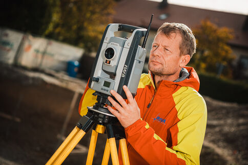 Ein Mann in einer orangegelben Jacke bedient ein Totalstation-Vermessungsgerät auf einem Stativ im Freien; im Hintergrund sind Gebäude und Bäume zu sehen.