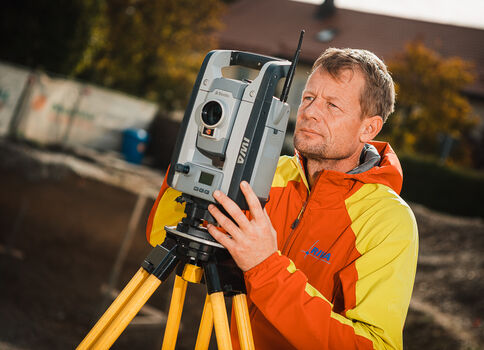 Ein Mann in einer orangegelben Jacke bedient ein Totalstation-Vermessungsgerät auf einem Stativ im Freien; im Hintergrund sind Gebäude und Bäume zu sehen.