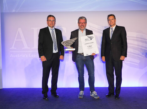 Drei Männer stehen auf einer Bühne bei einer Preisverleihung. Der mittlere Mann hält einen Pokal und eine Urkunde, während die beiden anderen in Anzügen daneben stehen und lächeln.