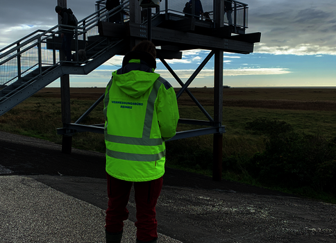Eine Person in einer neon-grünen Sicherheitsweste und roter Hose steht mit dem Rücken zur Kamera auf einer asphaltierten Fläche. Vor ihr erhebt sich eine erhöhte Metall-Aussichtsplattform mit Treppen und Geländern; darauf stehen mehrere Menschen und blicken auf eine weite, flache Landschaft bei wolkigem Himmel.