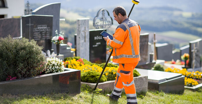 A maintenance worker in high-visibility orange clothing trims grass with a long-handled tool in a cemetery, surrounded by gravestones and flowers, with a rural landscape in the background.
