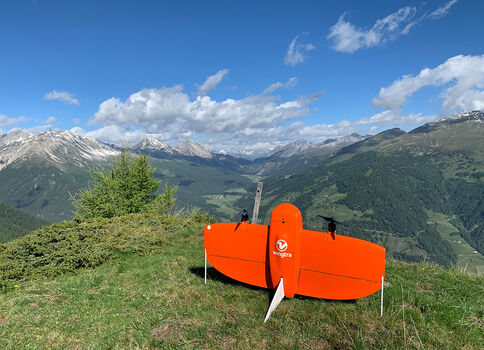 Auf einer grasbewachsenen Bergwiese steht ein leuchtend oranges Kleinfuggerät; im Hintergrund erstrecken sich grüne Täler und schneebedeckte Gipfel unter einem blauen Himmel mit Wolken.