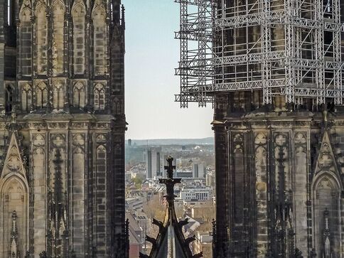 Zwei gotische Kirchtürme stehen eng beieinander: links reich verziert, rechts von Gerüsten bedeckt. Zwischen ihnen ragt ein spitzer Turm mit Kreuz empor; im Hintergrund eine städtische Skyline. Die Kathedrale scheint derzeit renoviert zu werden.
