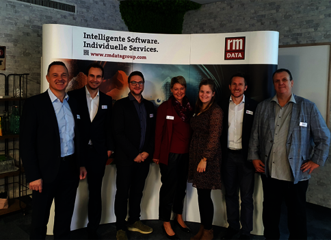 Sieben Personen in Business-Outfits stehen nebeneinander vor einem Messestand mit Banner "Intelligente Software. Individuelle Services." und dem rm DATA-Logo; alle tragen Namensschilder und lächeln.