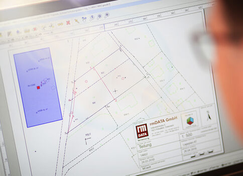 Eine Person schaut auf einen Computerbildschirm mit einem technischen Plan oder Grundriss: Geometrische Linien, Grundstücksgrenzen, ein großes blaues Rechteck links und mehrere beschriftete Felder.