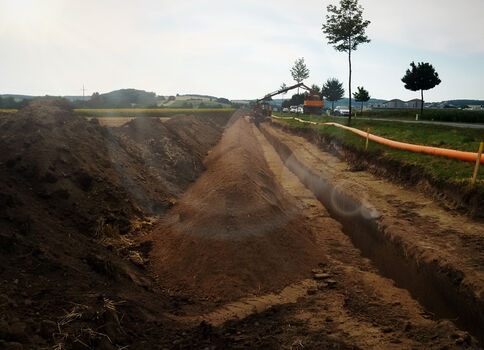 Baustelle auf dem Land: tiefe Gräben mit aufgeschüttetem Erdreich, eine orangefarbene Rohrleitung schlängelt sich rechts entlang. Hintergrund Felder, einzelne Bäume und Baufahrzeuge unter hellem Himmel.