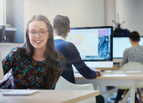 Eine junge Frau mit roten Brillenrahmen sitzt lächelnd an einem hellen Arbeitsplatz in einem modernen Büro- oder Klassenraum und schaut in die Kamera. Vor ihr liegt ein Tablet oder Notizblock; im Hintergrund arbeiten weitere Personen an Computerbildschirmen.