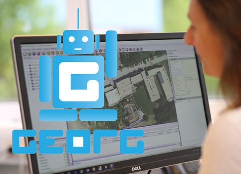 Eine Person sitzt vor einem Computerbildschirm und betrachtet eine Geodaten-/Kartenansicht, vermutlich Satelliten- oder Luftbildaufnahmen.