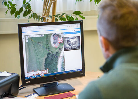 Eine Person sitzt von hinten an einem Schreibtisch und betrachtet am Computer eine Kartenansicht mit Satellitenbild auf dem Monitor.