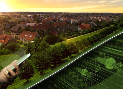 Eine Person hält ein Smartphone; im Hintergrund erstreckt sich eine Vorstadtlandschaft bei Sonnenuntergang. Rechts leuchtet eine grüne digitale Netzdarstellung auf einer Glasfläche und symbolisiert vernetzte Städte.
