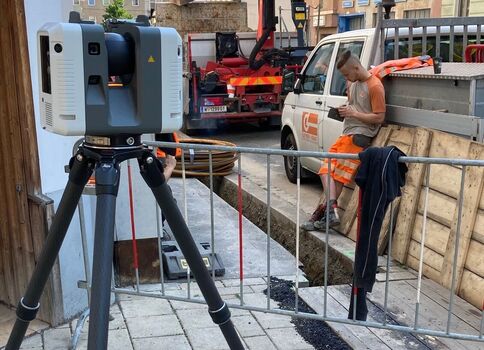 Vermessungsgerät auf Stativ vorne, Baustelle in der Stadt; Absperrungen, Bauarbeiter in Orange hinter dem Geländer, ein Baufahrzeug mit Ausleger in der Straße, Häuser im Hintergrund.