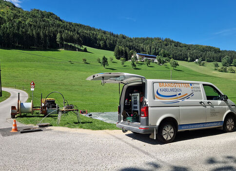 Auf einer sonnigen Landstraße steht ein silberner Servicewagen mit offener Heckklappe; daneben liegt ein Anhänger mit Ausrüstung und eine orangefarbene Warnstange. Im Hintergrund erstrecken sich grüne Wiesen, Wälder und sanfte Hügel mit Häusern.