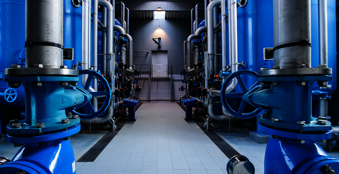 Industrial facility with two parallel rows of large blue pipes and valves on either side, forming a central aisle that leads to a doorway. A metal floor and overhead lighting frame the symmetrical machinery, suggesting a pumping or processing plant.
