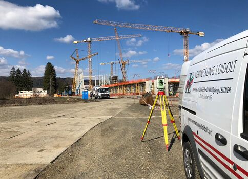 Eine Großbaustelle mit mehreren Krananlagen und Baugerüsten. Im Vordergrund steht ein Vermessungsgerät auf einem Stativ; daneben ein weißer Service-Van.