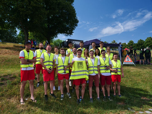 Gruppe junger Menschen in leuchtend gelben Sicherheitswesten und roten Shorts posiert auf einer grasigen Wiese bei sonnigem Himmel; im Hintergrund stehen Bäume und ein Veranstaltungsbereich.