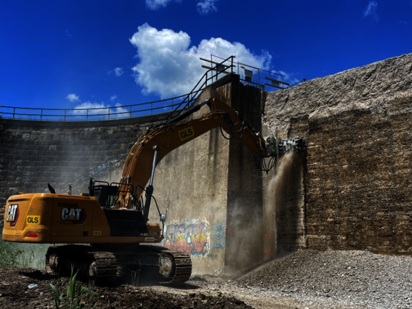 Bright yellow CAT excavator with a long hydraulic arm demolishes material inside a circular brick wall, lifting debris as dust rises. A clear blue sky with clouds overlooks the scene.