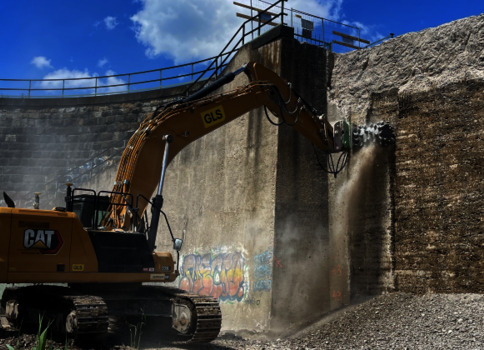 Ein gelber CAT-Bagger arbeitet an einer steinernen Ufermauer und gräbt Erde aus dem Bereich vor der Mauer. Kies und Geröll liegen auf dem Boden; der Himmel ist klar blau mit einigen Wolken.